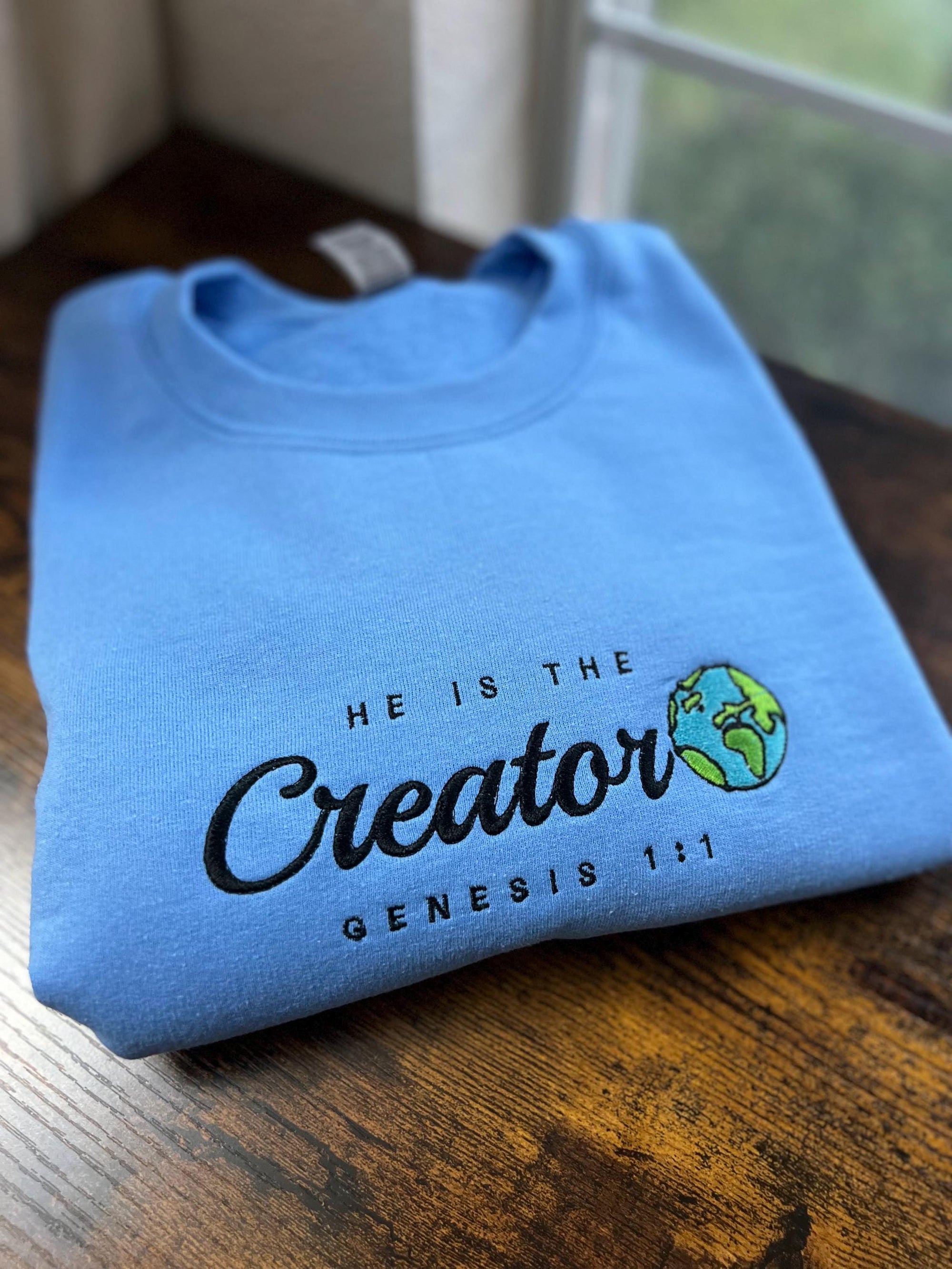 A blue crewneck sweatshirt with the text 'HE IS THE Creator GENESIS 1:1' embroidered on the front.