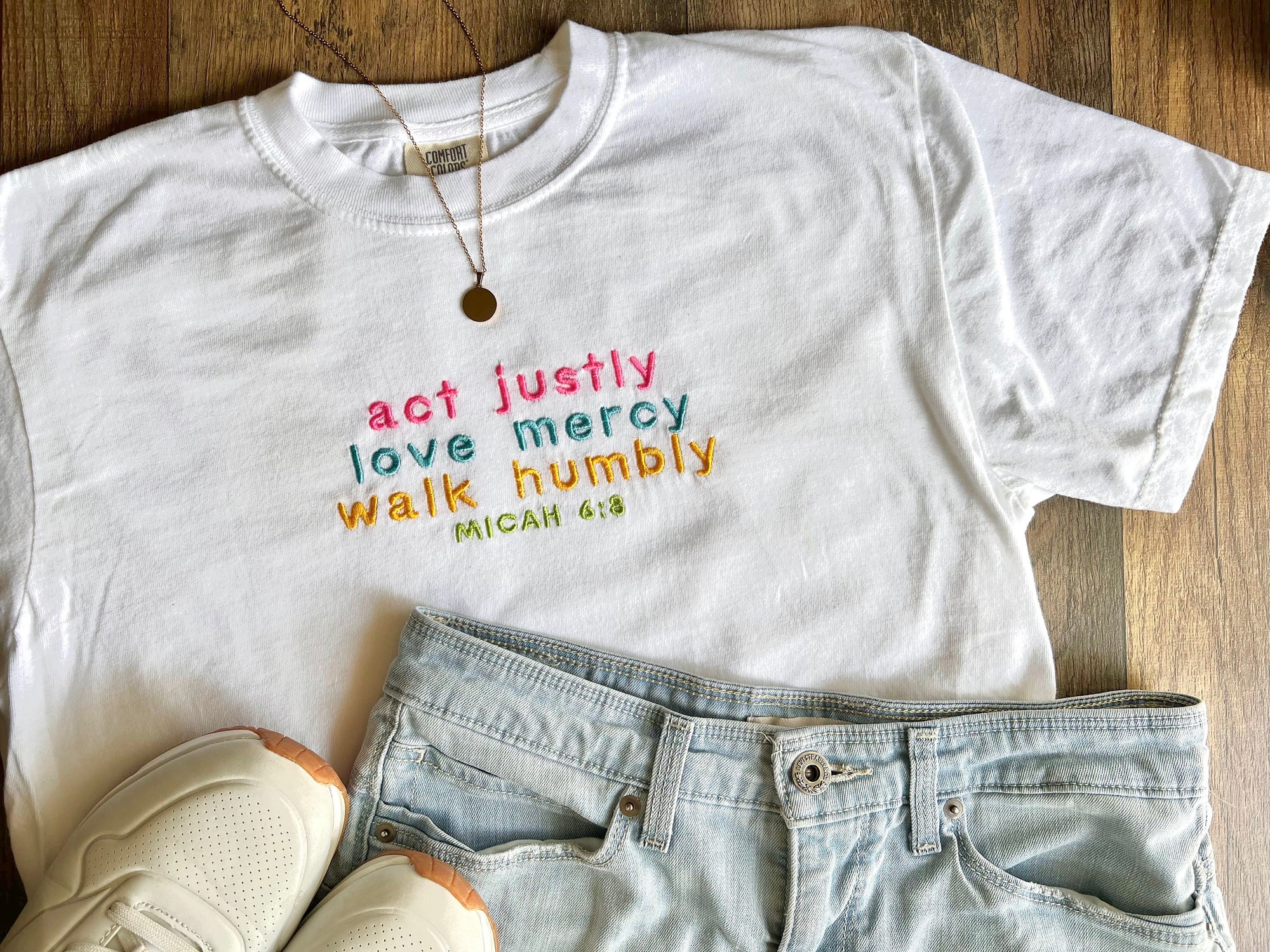 White embroidered t-shirt with the text 'act justly, love mercy, walk humbly, MICAH 6:8' laid flat with a pair of blue jeans and white sneakers.