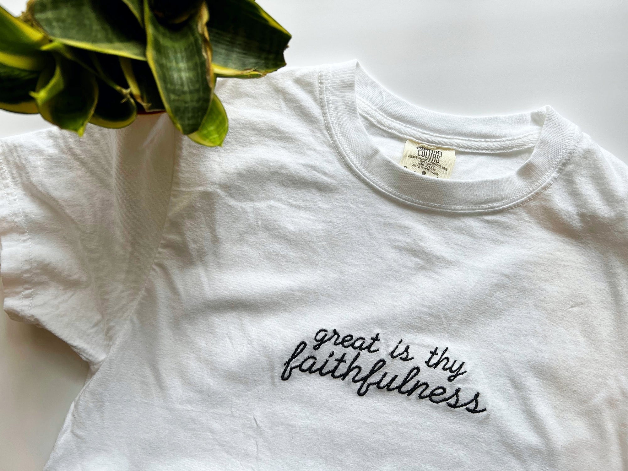 White crew neck t-shirt with the text 'Great is Thy Faithfulness' embroidered in black on the chest, displayed flat with a green plant in the background.