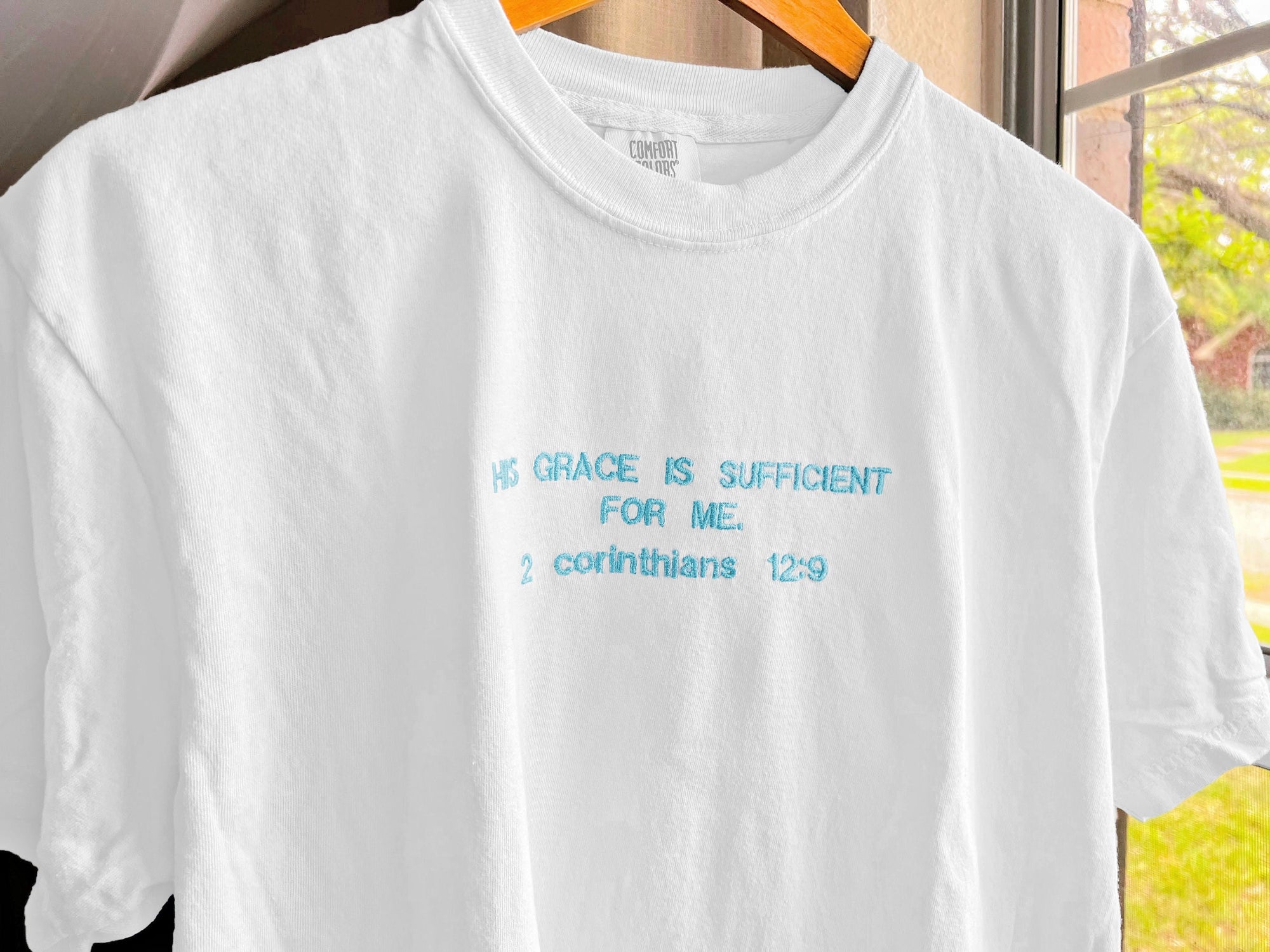 White embroidered t-shirt with the Bible verse 'His grace is sufficient for me' in blue text.