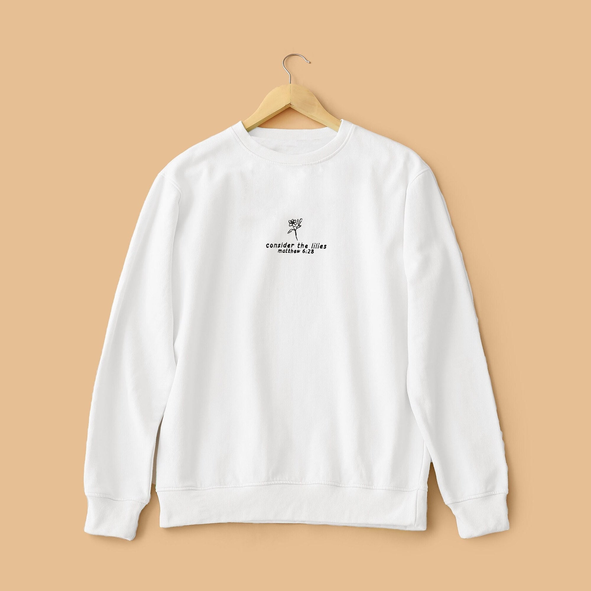White crewneck sweatshirt with text embroidery, hanging against a beige background.