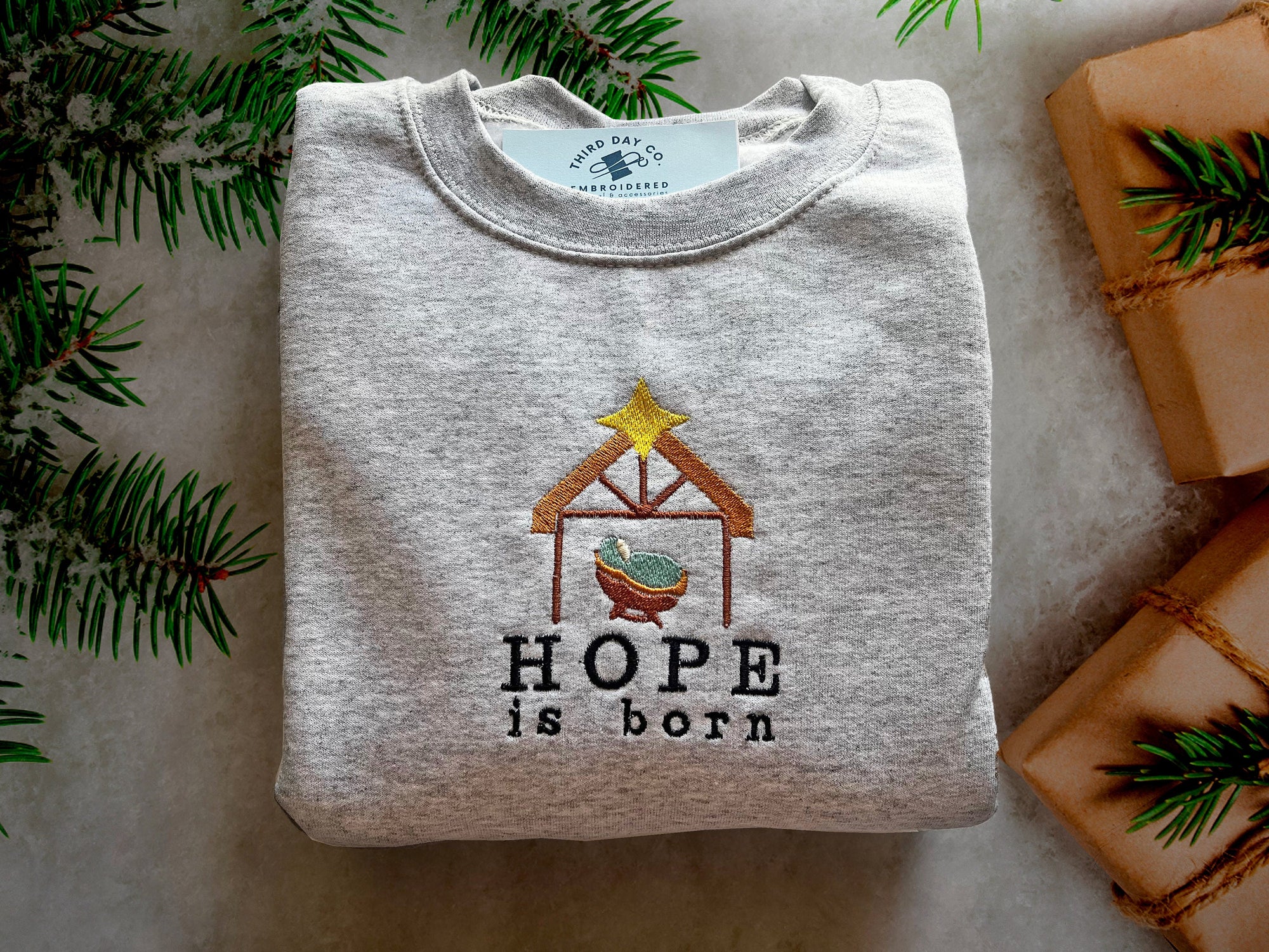 A gray sweatshirt with the phrase 'HOPE is born' embroidered on the front, featuring a small nativity scene.