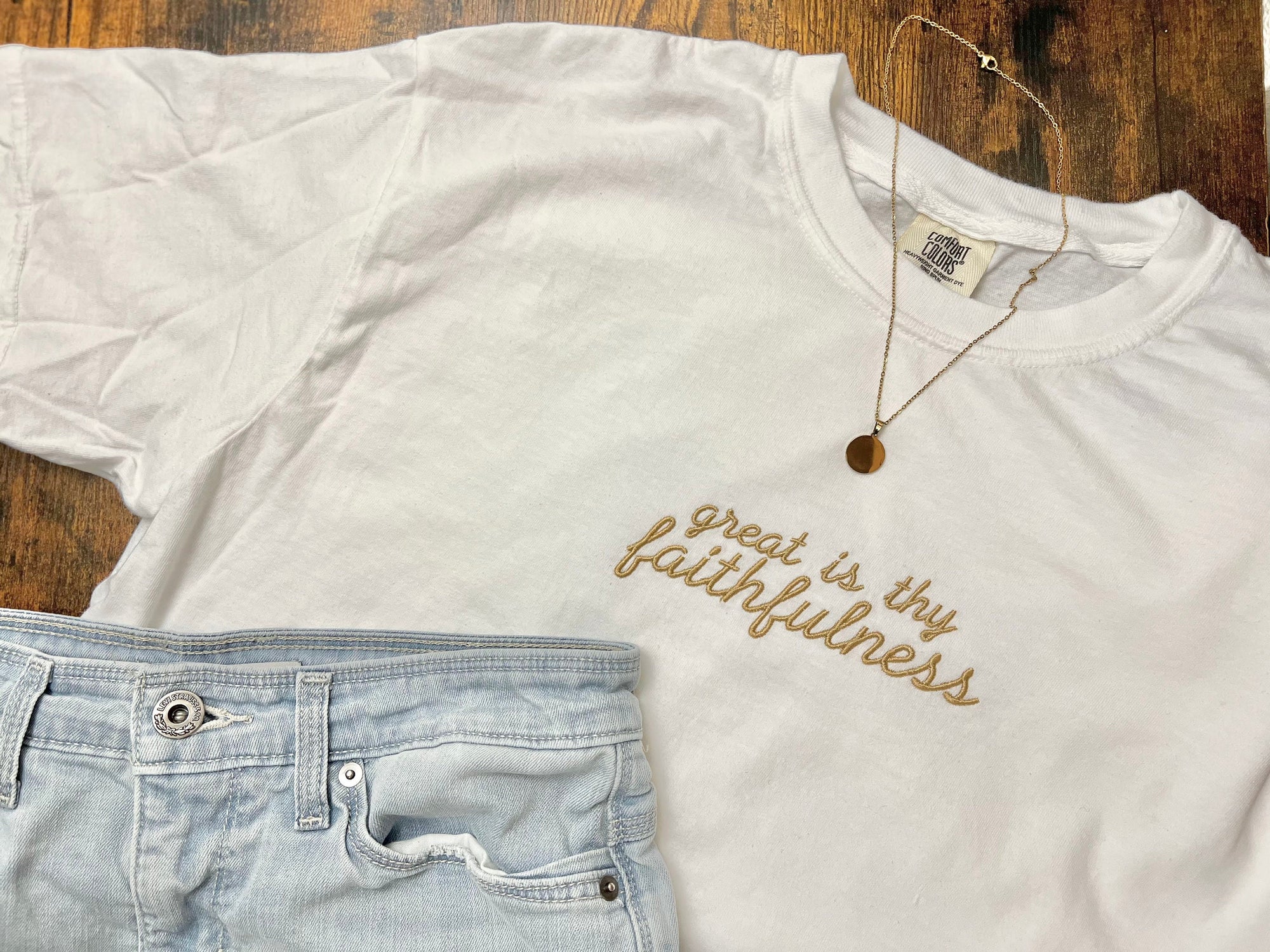 White embroidered t-shirt with the phrase 'Great is Thy Faithfulness' laid out flat alongside light blue jeans and a necklace, all placed on a wooden surface.