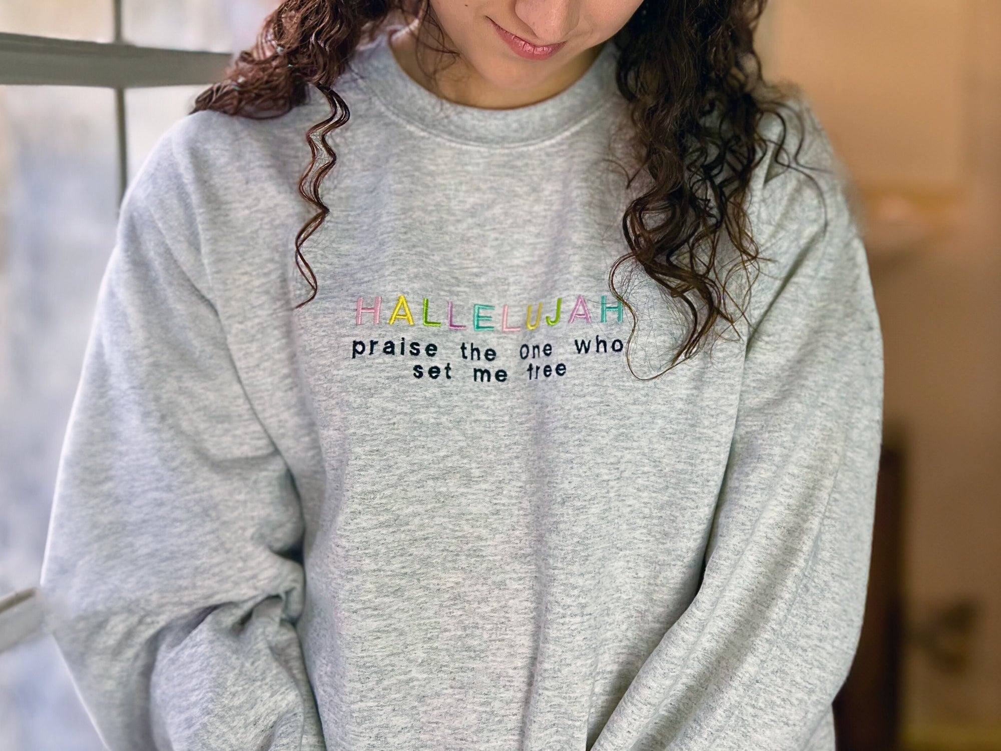 A person wearing an ash gray crewneck sweatshirt with the embroidered text 'Hallelujah praise the One who set me free' in a colorful font.