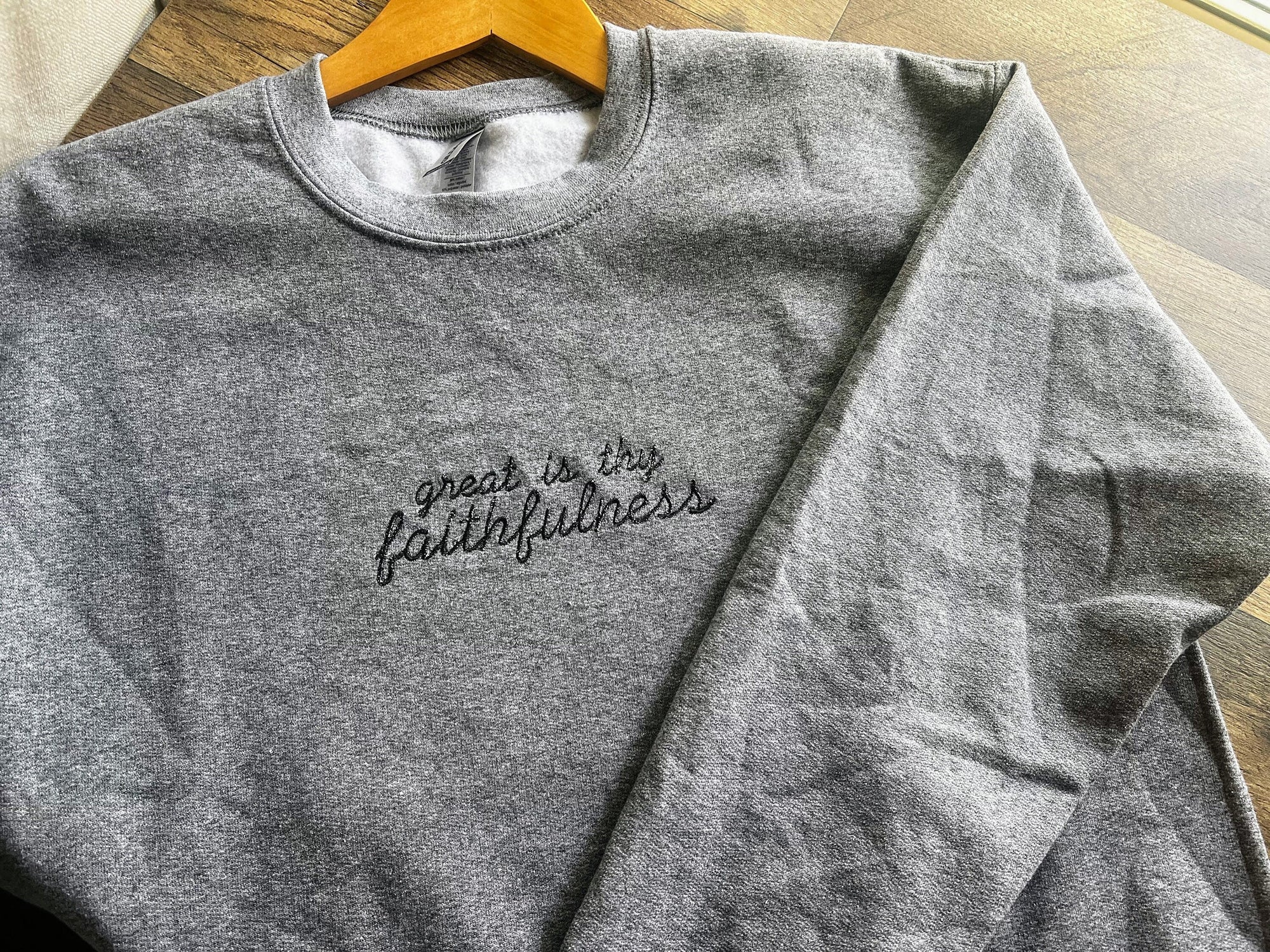 A gray embroidered sweatshirt with the text 'Great is Thy Faithfulness' displayed on a wooden hanger.