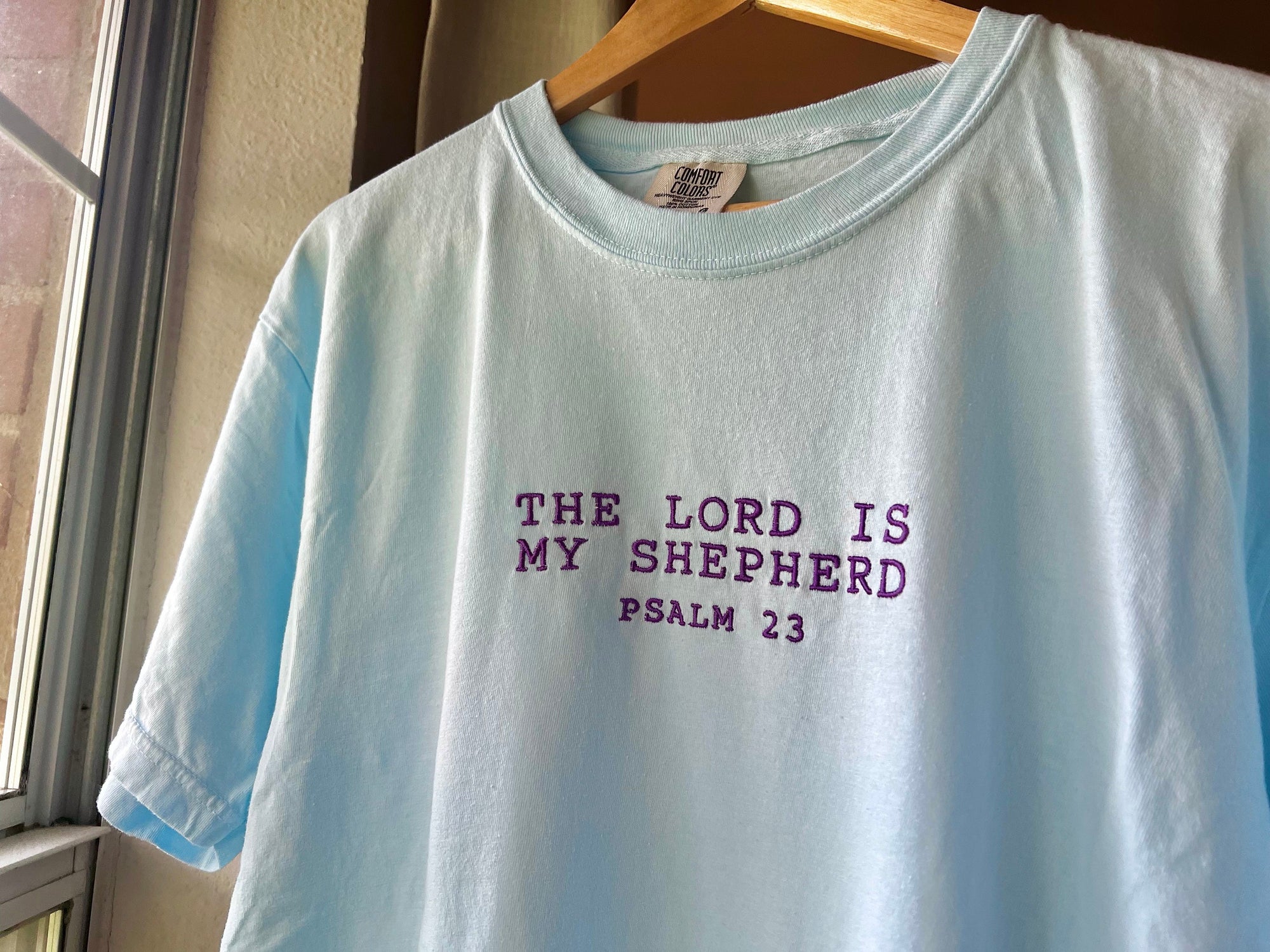 A blue embroidered crewneck t-shirt with 'THE LORD IS MY SHEPHERD PSALM 23' written in purple embroidery.