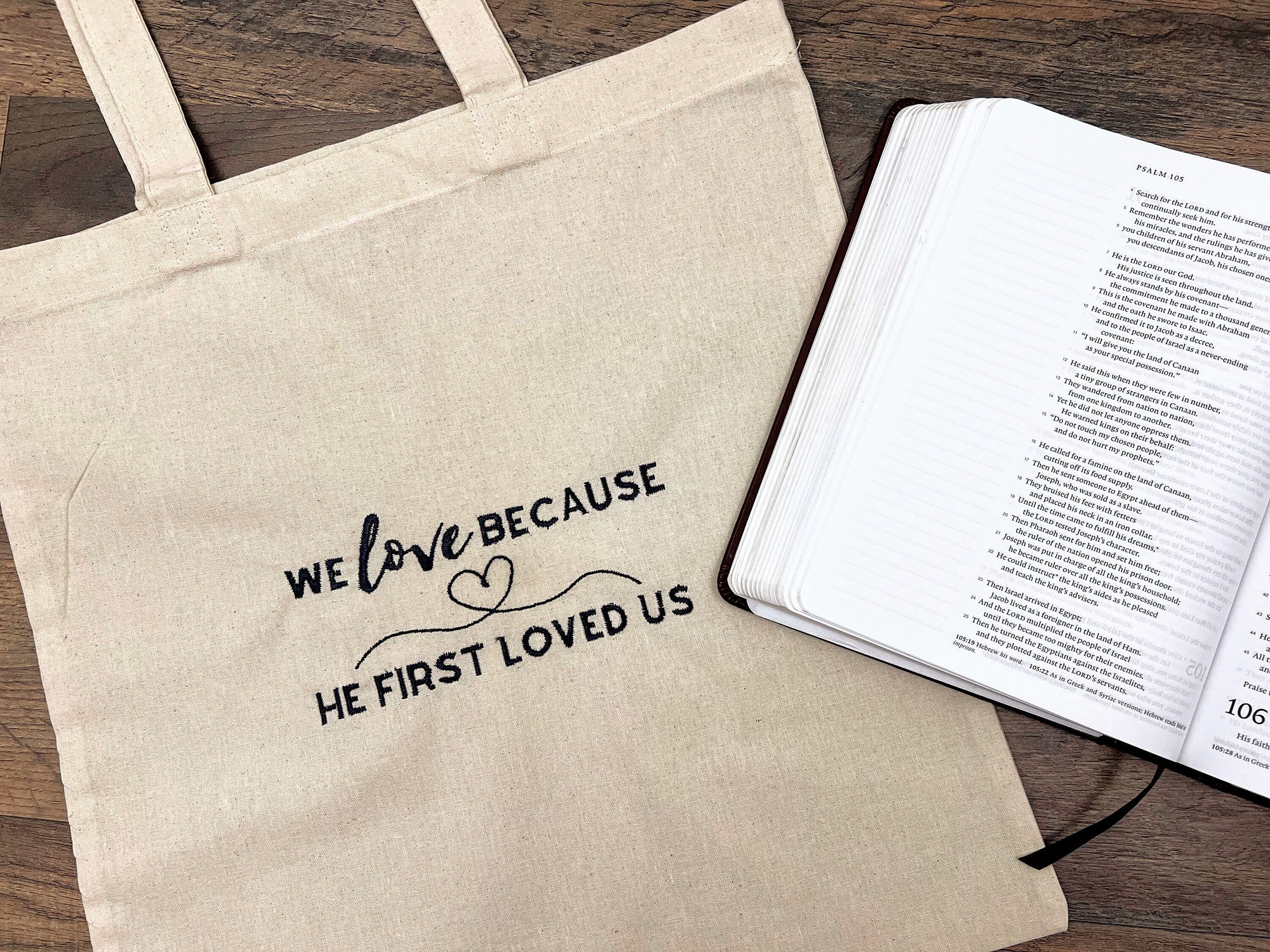 A beige canvas tote bag with the embroidered text 'WE LOVE BECAUSE HE FIRST LOVED US' alongside an open Bible on a wooden surface.