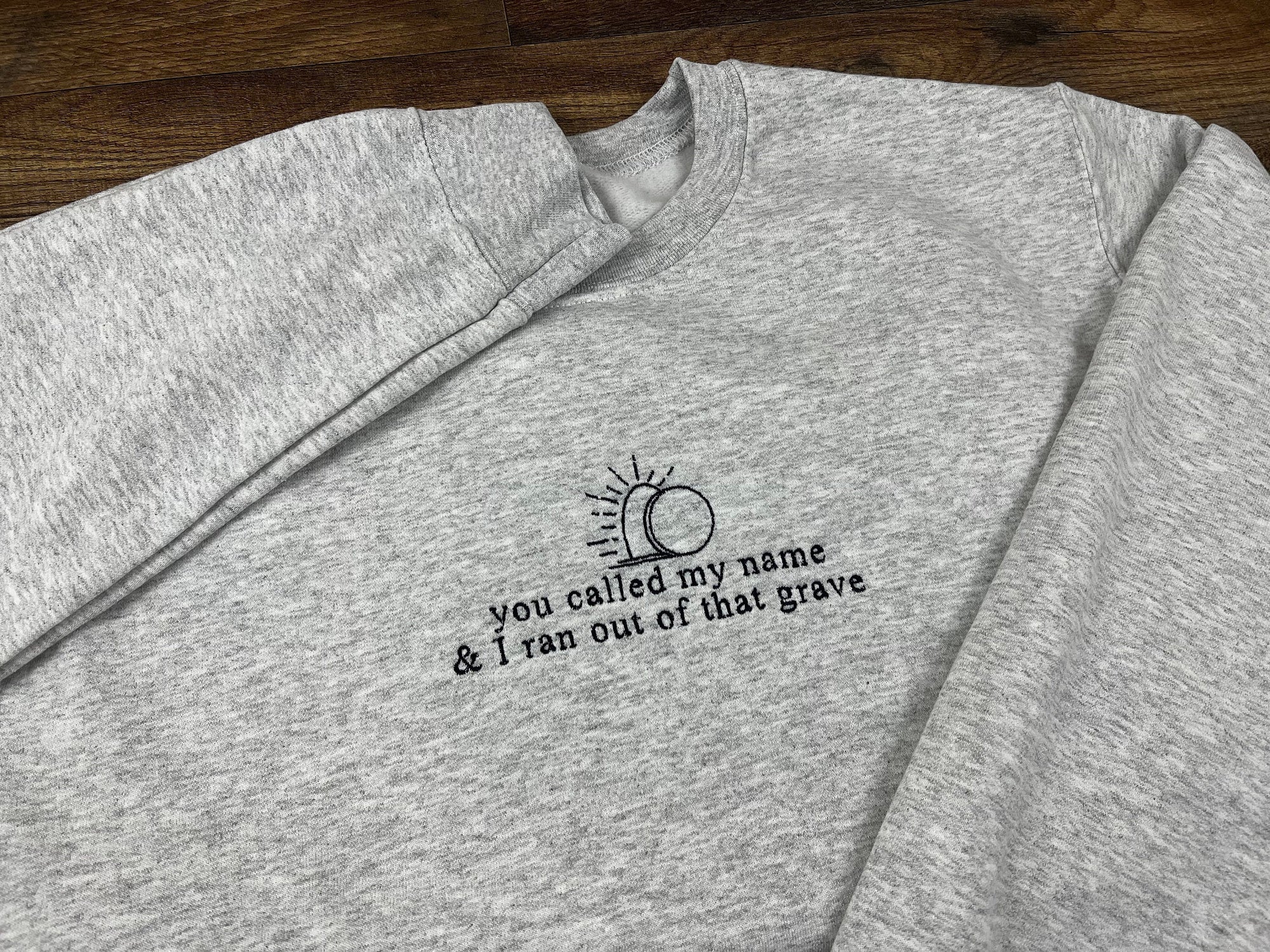 A gray sweatshirt with dark navy blue embroidered text and design, featuring a crewneck style.
