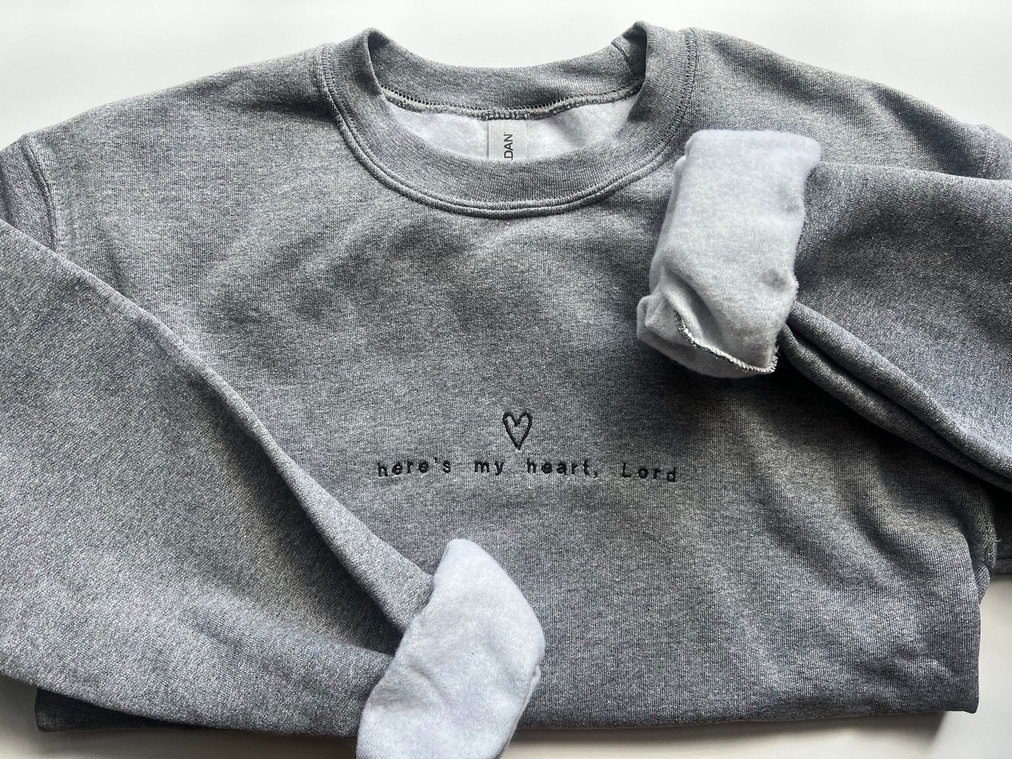 Heathered gray sweatshirt with embroidered text 'Here's my heart, Lord' in black lettering, displayed flat with sleeves folded.
