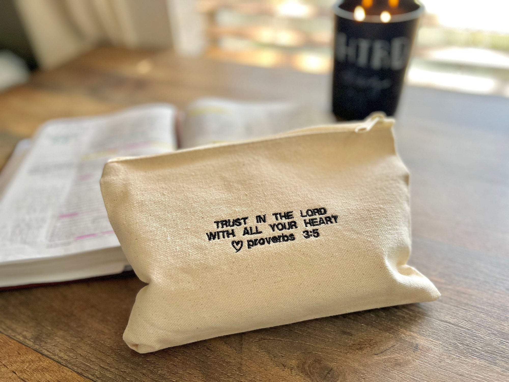 A beige canvas zipper pouch with the embroidered text 'TRUST IN THE LORD WITH ALL YOUR HEART Proverbs 3:5' on the front, placed on a table beside a candle and an open Bible.