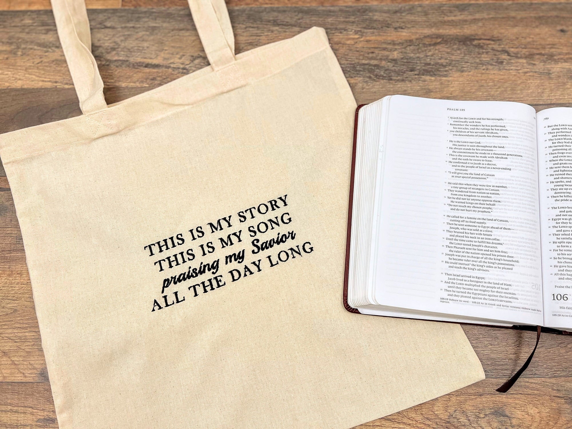 A beige canvas tote bag with black text embroidered that reads 'This is my story, this is my song Praising my Savior all the day long', displayed next to an open Bible.