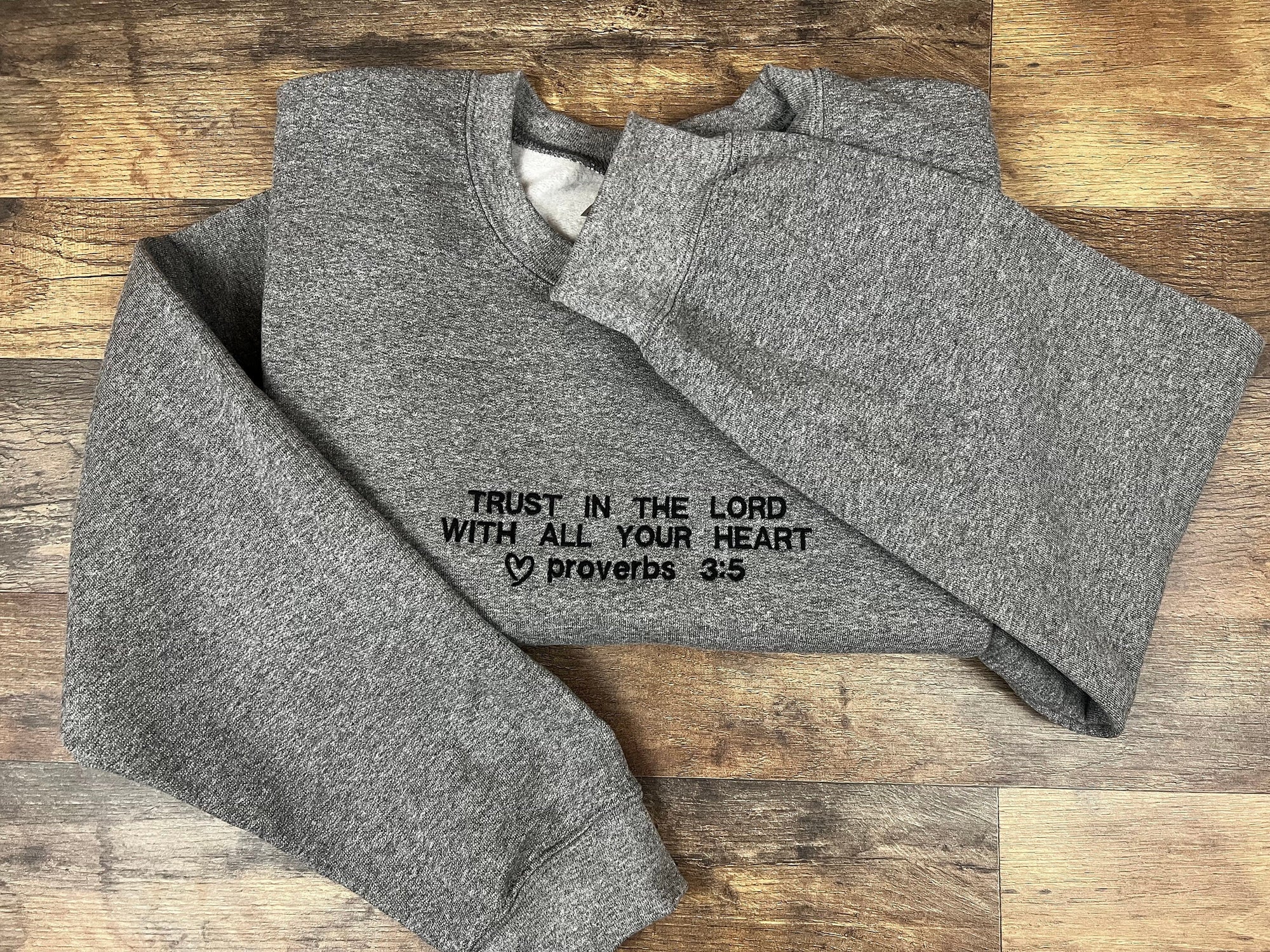 A gray embroidered crewneck sweatshirt with the text 'TRUST IN THE LORD WITH ALL YOUR HEART Proverbs 3:5' in black.