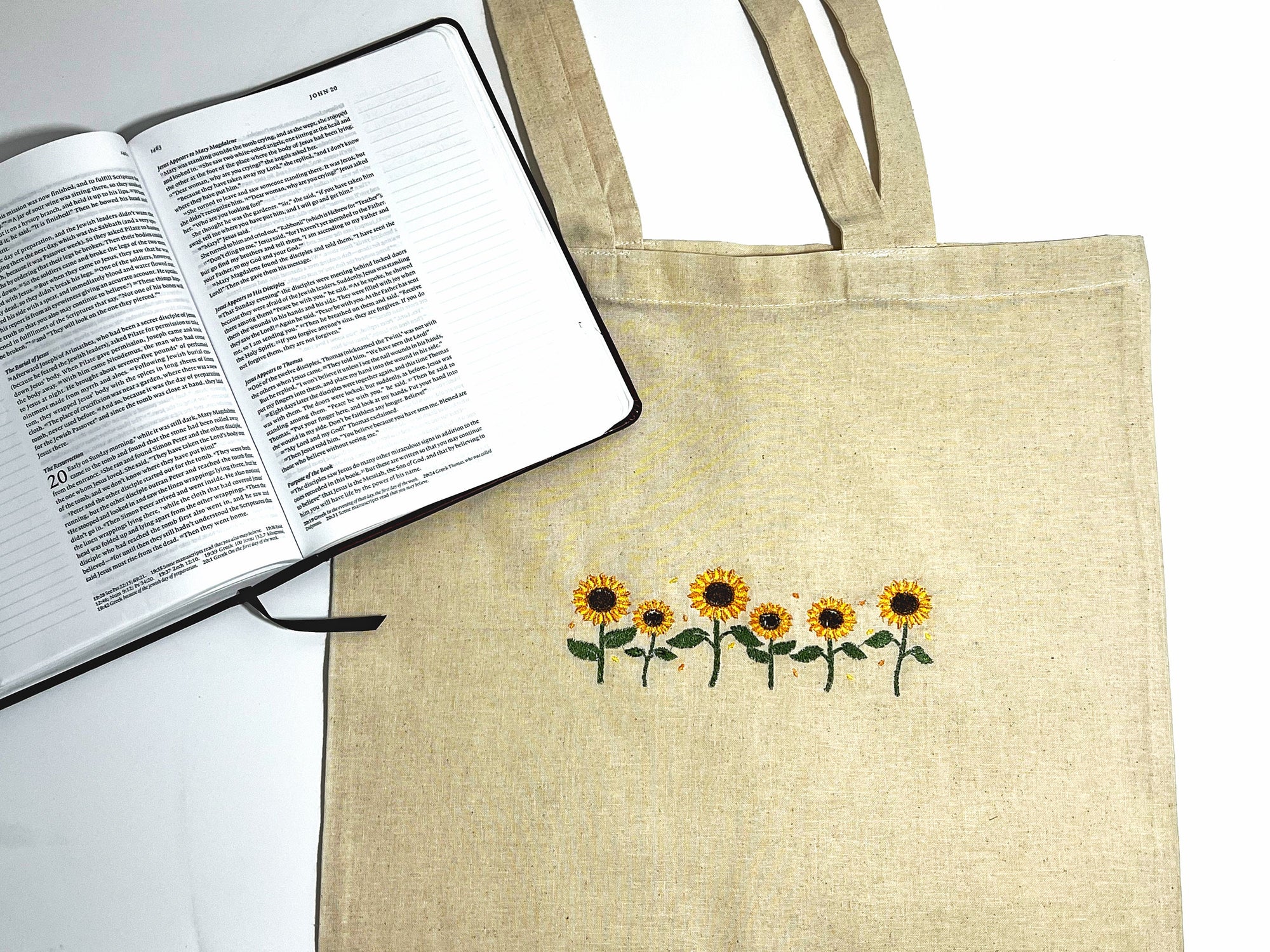 A beige canvas tote bag with embroidered yellow sunflowers on the front, displayed next to an open book.