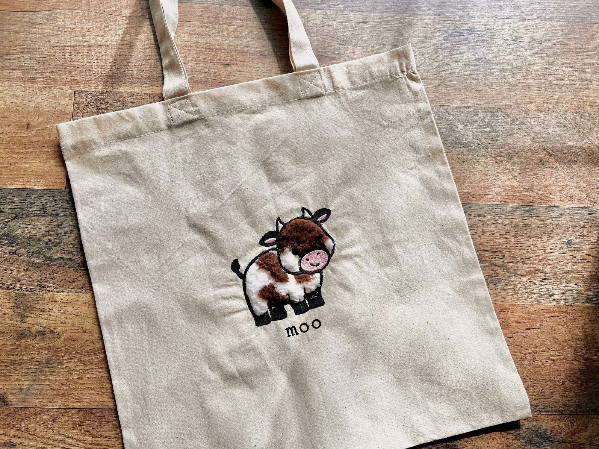 A beige canvas tote bag with an embroidered design of a cute cow and the word 'moo' beneath it.