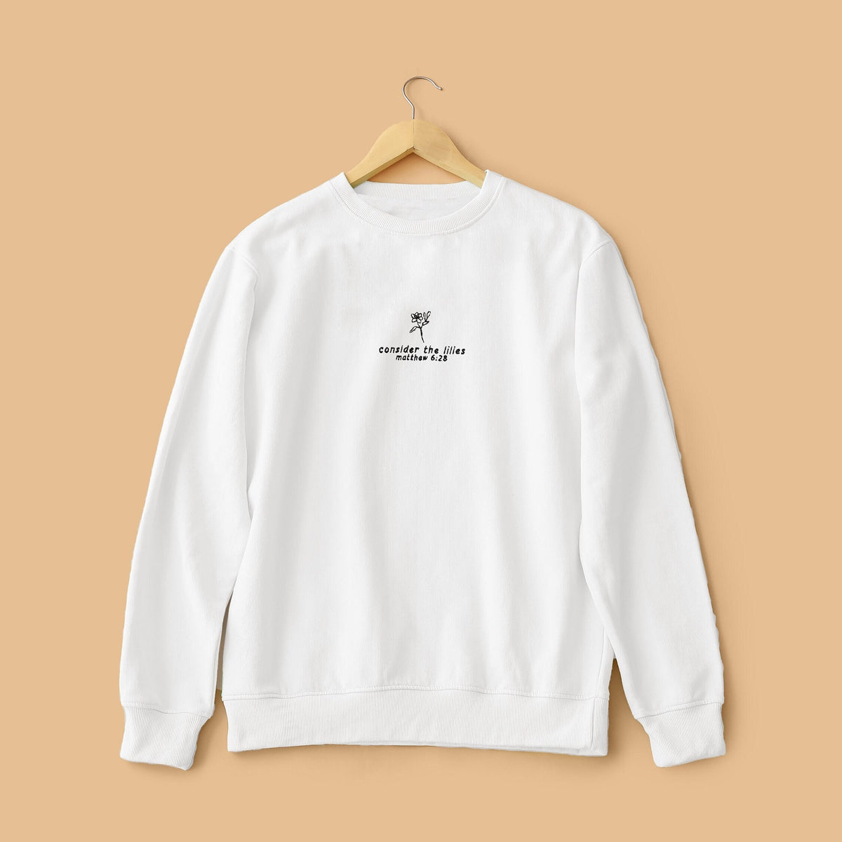 White crewneck sweatshirt with text embroidery, hanging against a beige background.