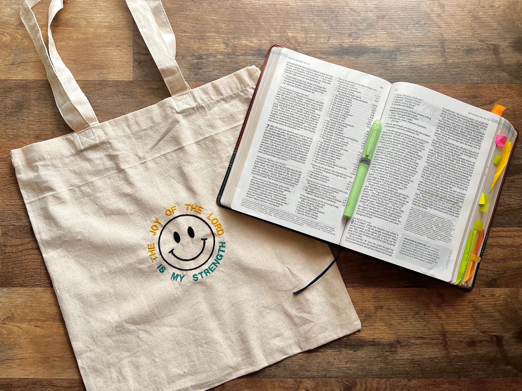 A beige canvas tote bag with a smiley face and a Bible verse embroidered on it, displayed next to an open Bible and a few writing instruments on a wooden surface.