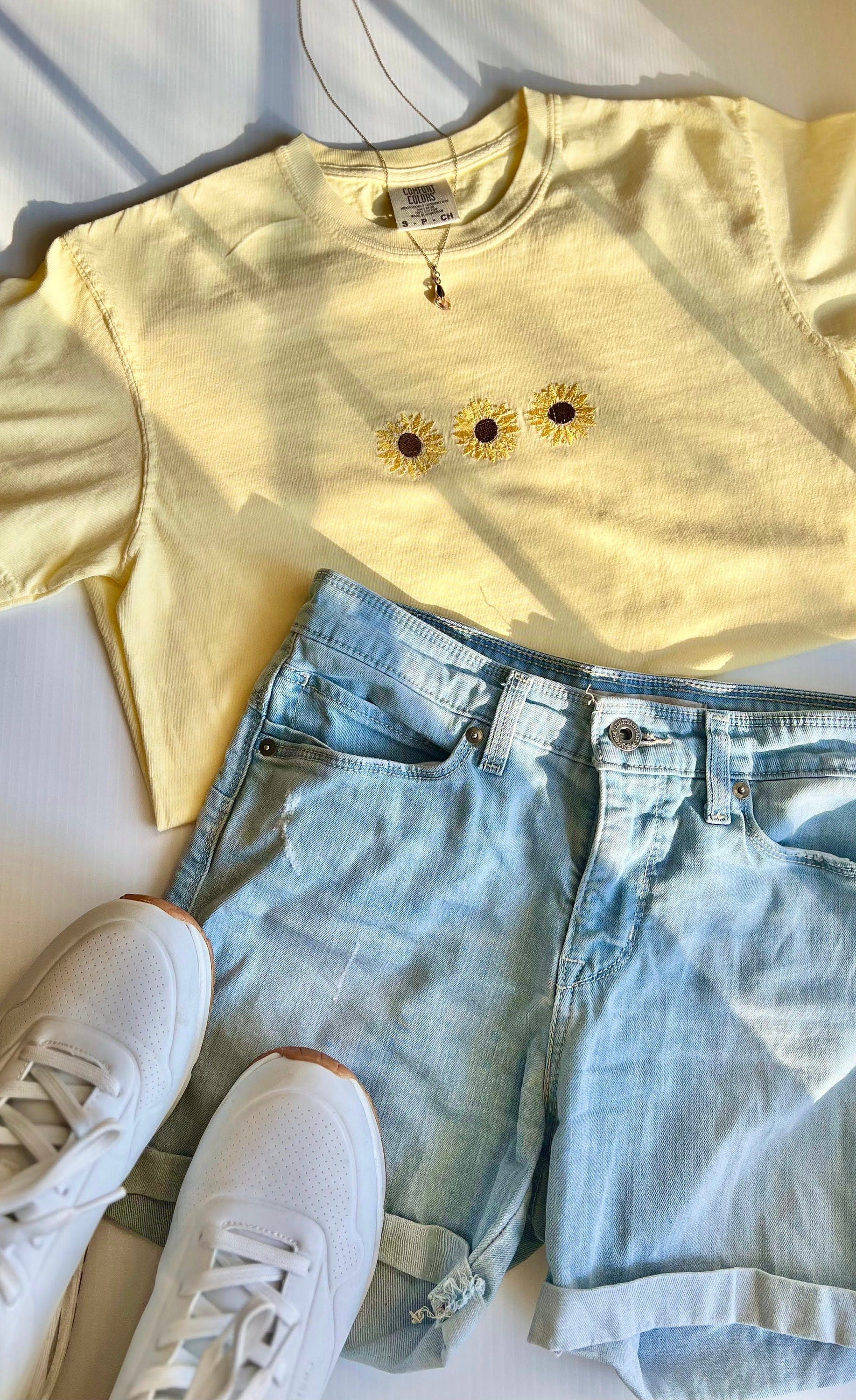 A yellow sunflower embroidered t-shirt laid flat with a pair of blue denim shorts and white sneakers.