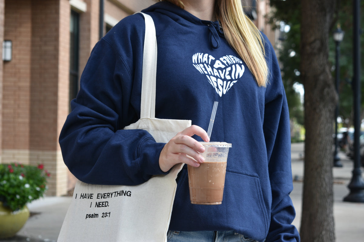 Person wearing a navy blue sweatshirt with text, holding a drink and a tote bag with text, outdoors.