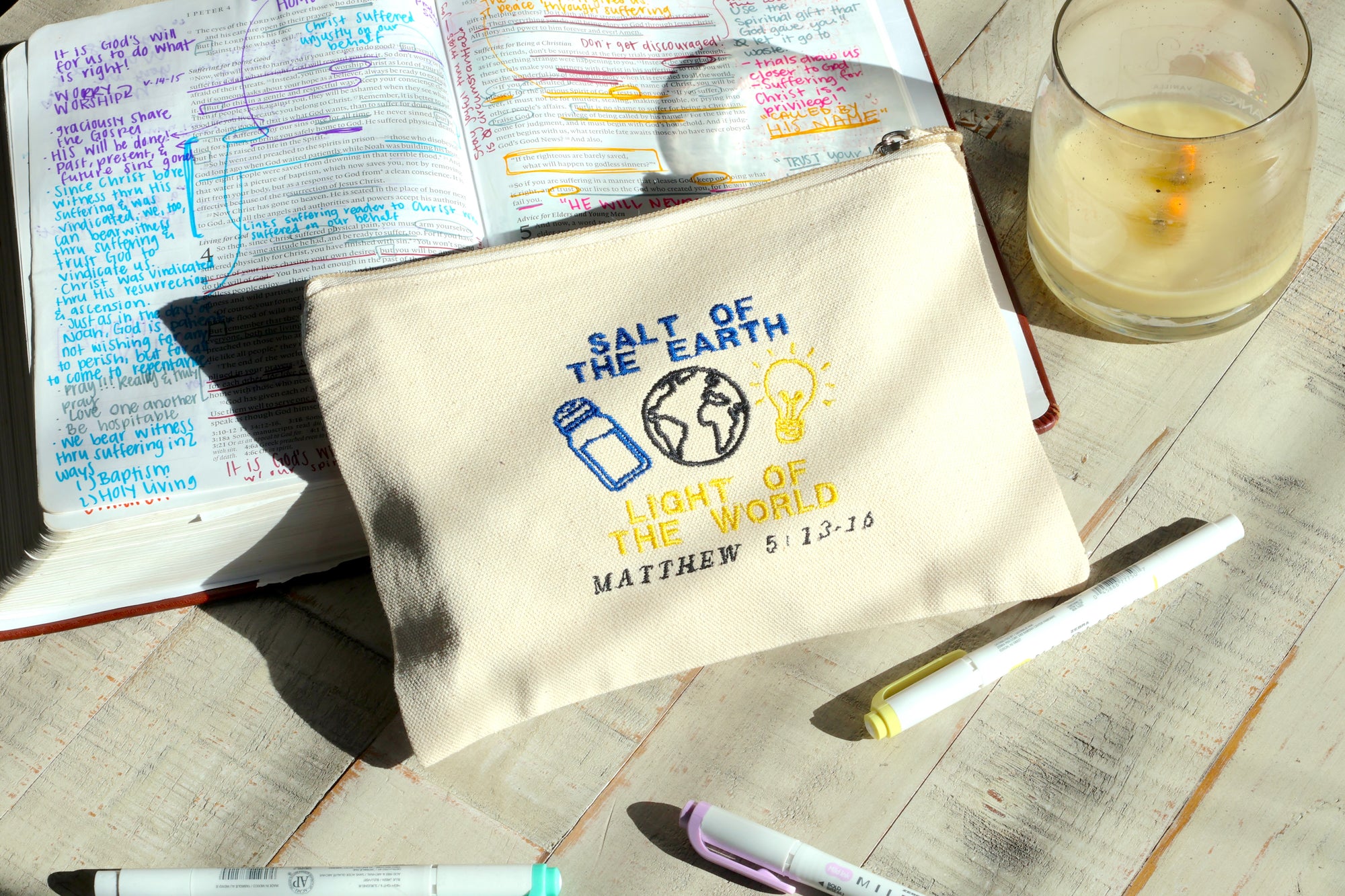 Canvas pouch with 'Salt of the Earth, Light of the World' text on a wooden surface with books and pens.