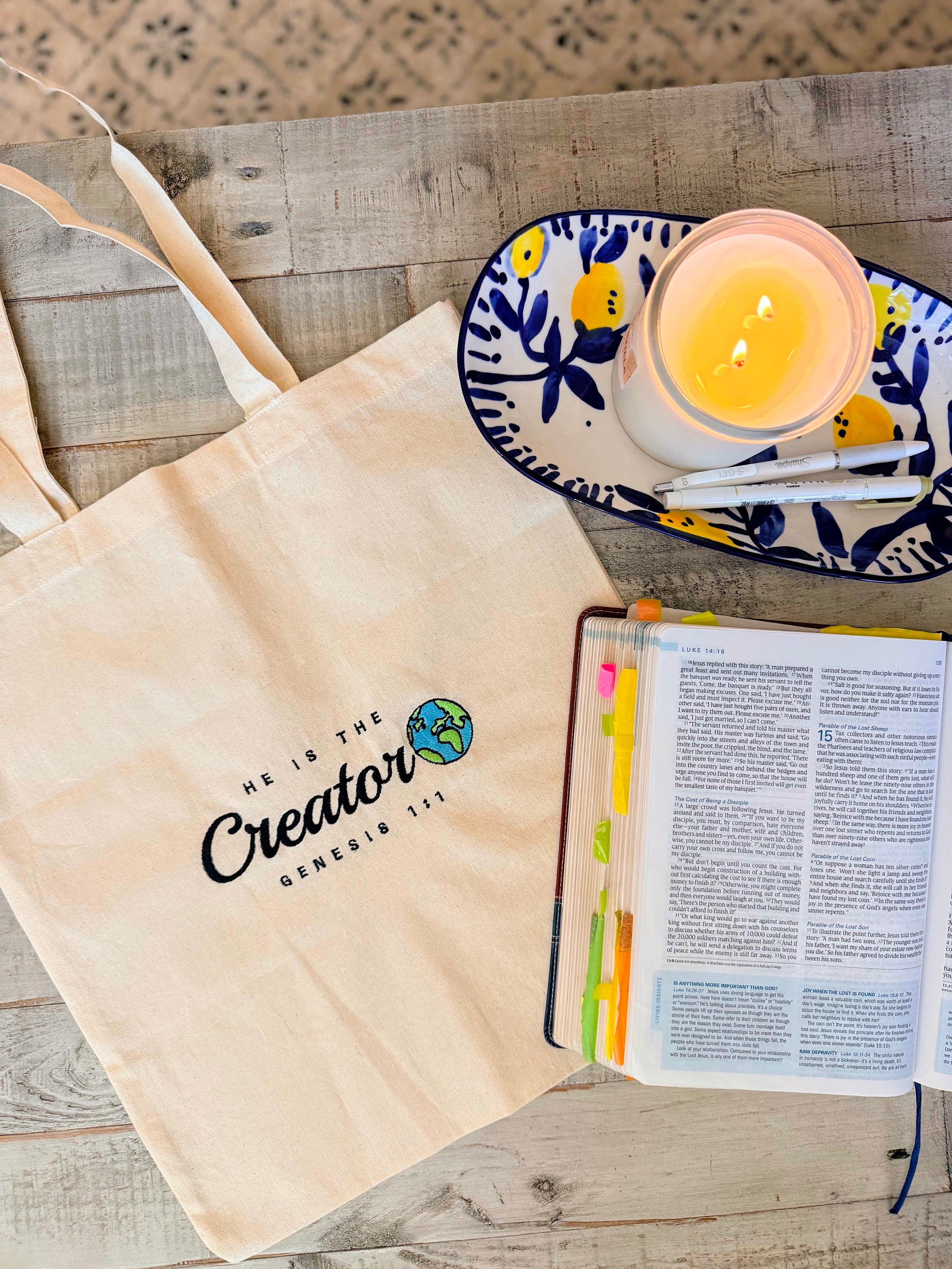 Tote bag with 'He is the Creator' text, open Bible, candle, and plate on a wooden surface