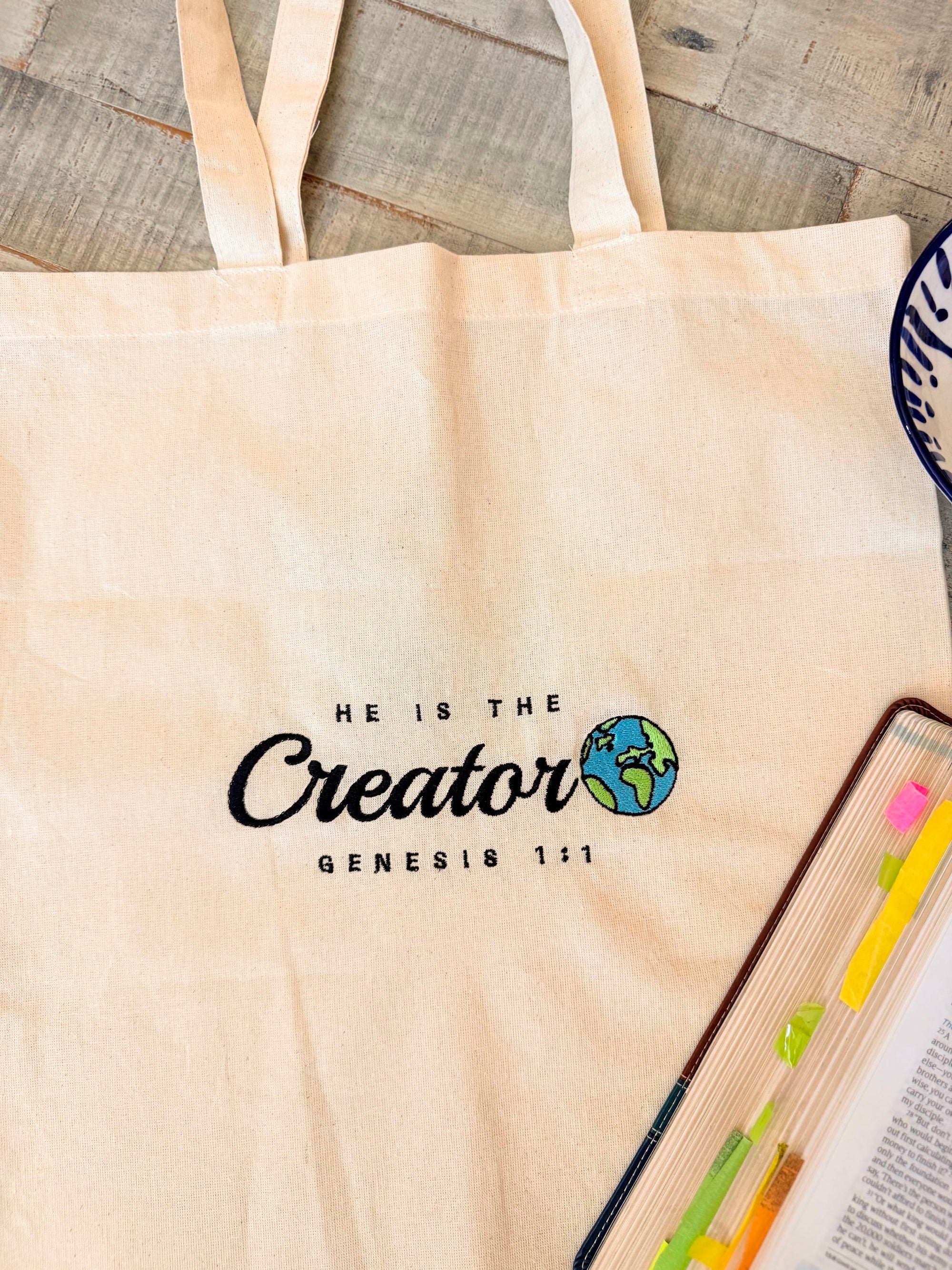 Beige tote bag with 'He is the Creator' text and globe graphic on a wooden surface.