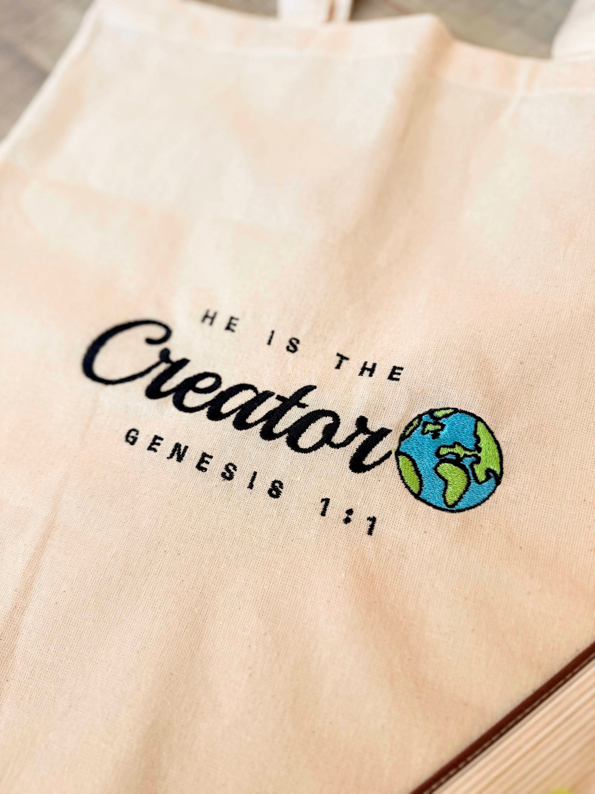 Beige tote bag with 'He is the Creator, Genesis 1:1' text and globe design.