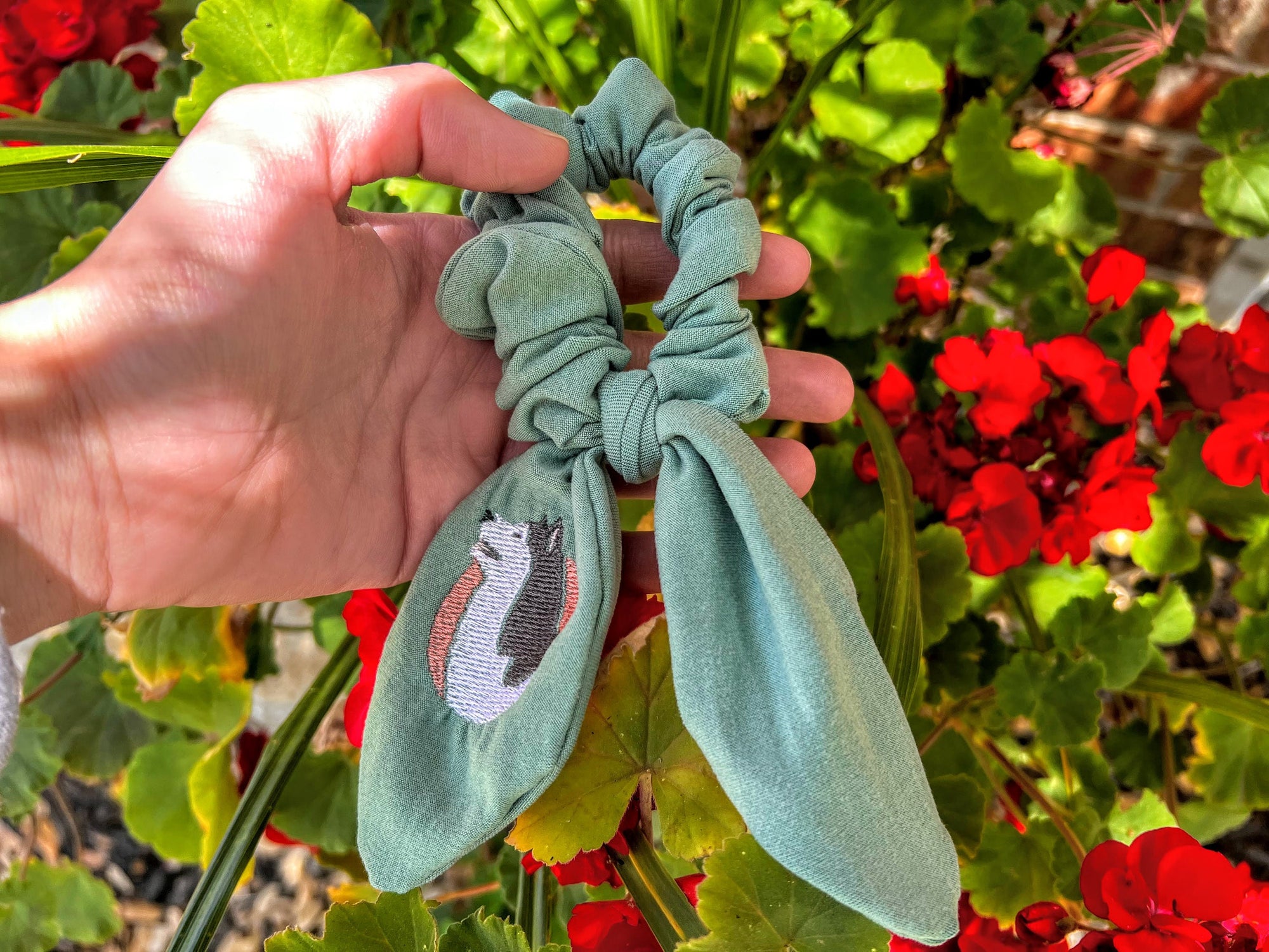 A hand holding a teal scrunchie with an embroidered husky design, with a background of green leaves and red flowers.