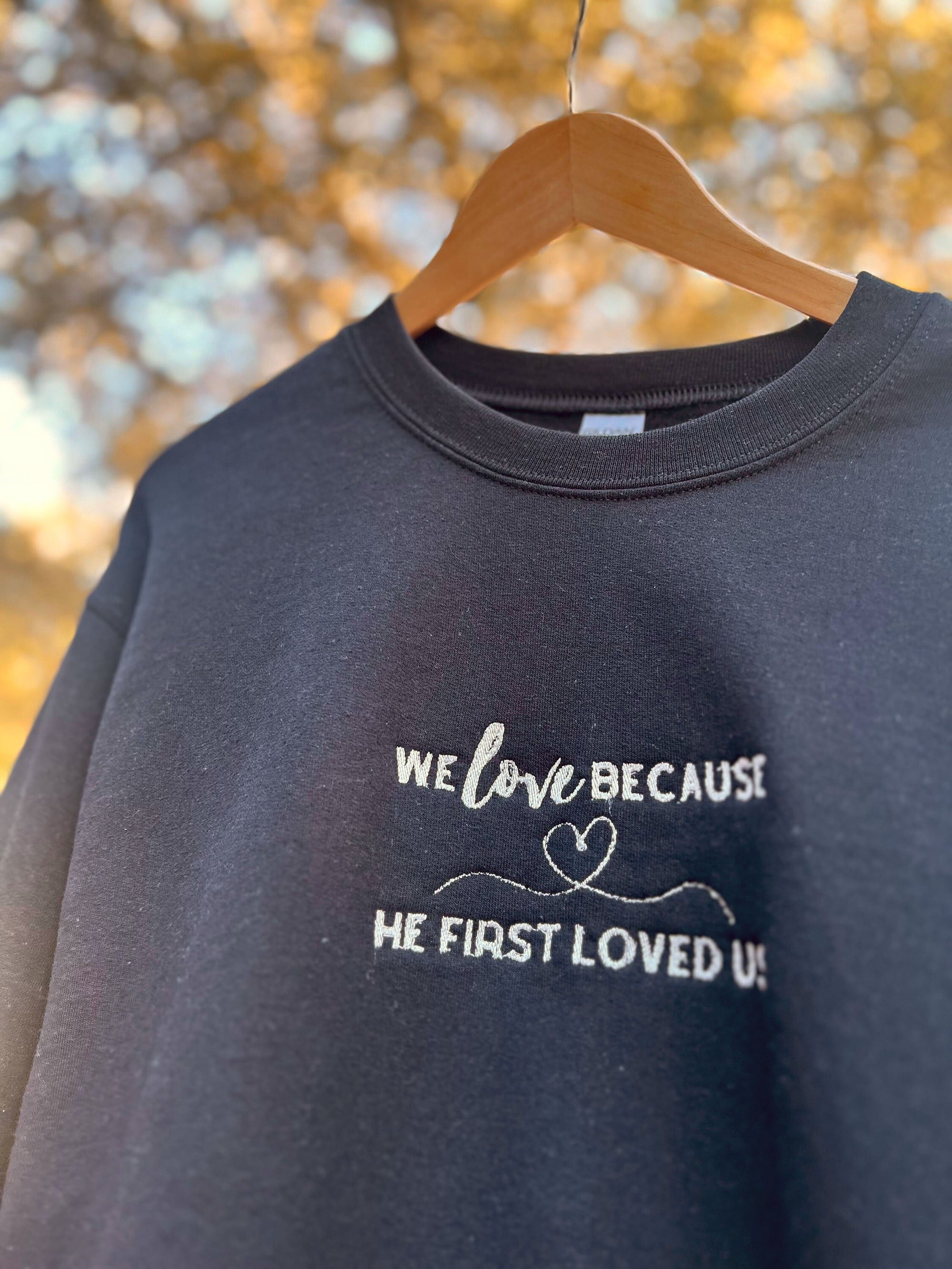 Black crewneck sweatshirt with the embroidered text 'WE LOVE BECAUSE HE FIRST LOVED US' on the front.