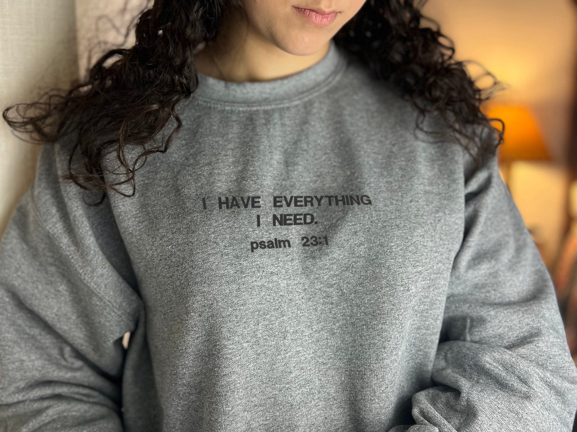 A person wearing a gray crewneck sweatshirt with the text 'I HAVE EVERYTHING I NEED. psalm 23:1' embroidered in black.