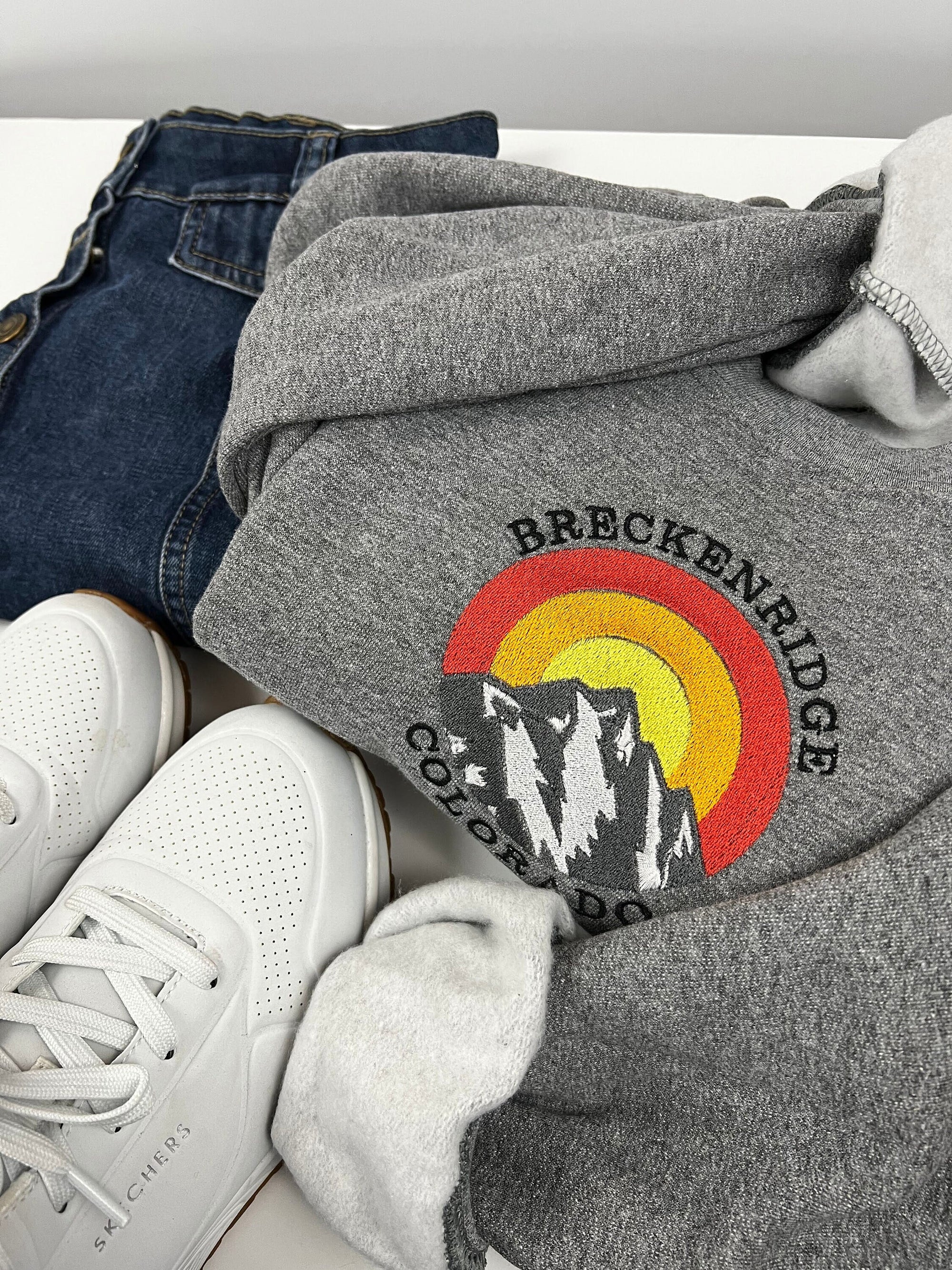 A gray crewneck sweatshirt with 'Breckenridge Colorado' embroidered on the front, displayed alongside a pair of shoes and a skirt.