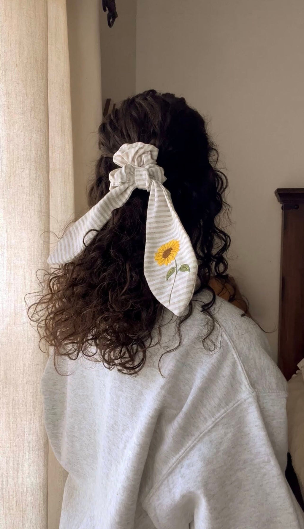A person with their back turned to the camera has their hair tied with a beige linen scrunchie that has an embroidered sunflower on it.